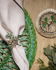 Decorative napkin ring with green leaves on a woven mat and wooden surface.