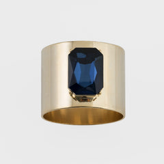 A gold-colored napkin ring with a navy blue gemstone centerpiece.