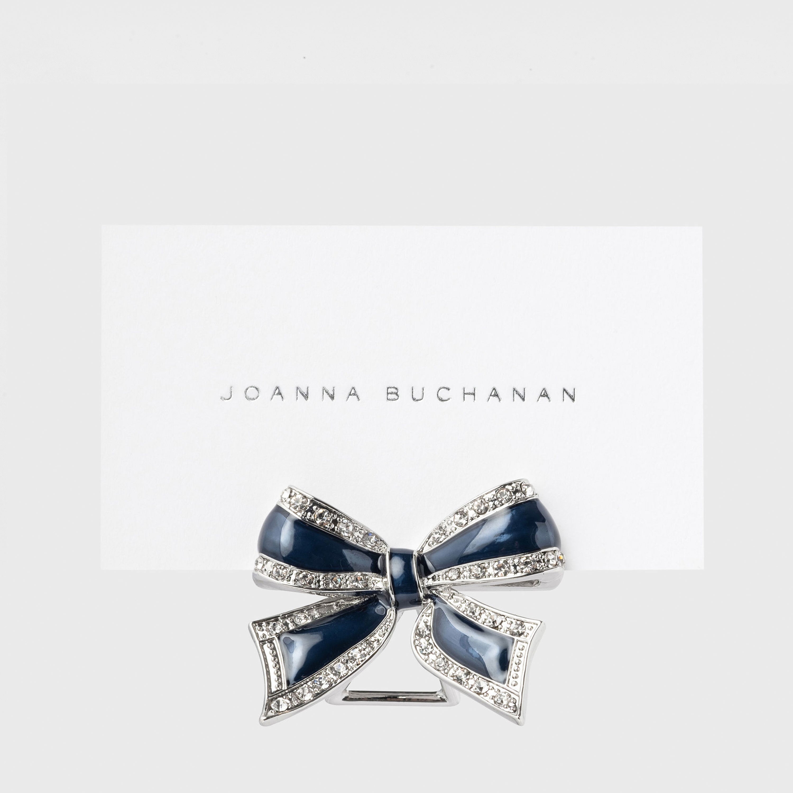 A navy and silver bow placecard holder with the brand name 'JOANNA BUCHANAN' written below it.