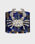 A blue and brown tortoiseshell resin napkin ring with a crab design in the center, adorned with sparkling embellishments.