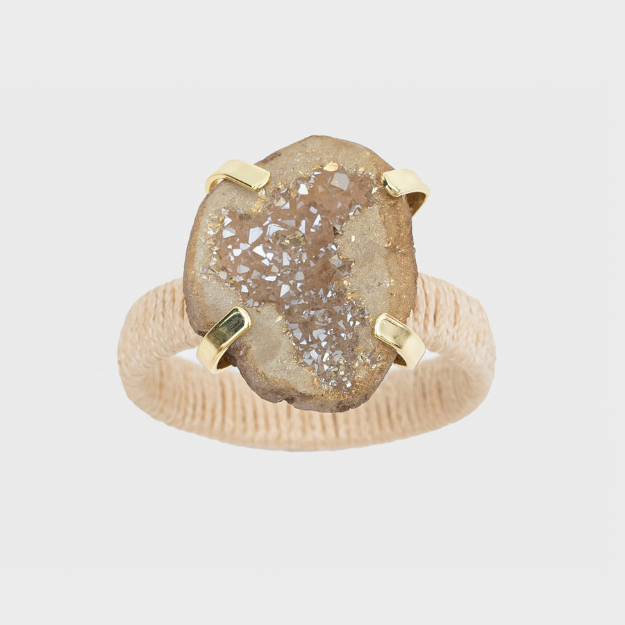 A napkin ring featuring a natural raffia band and a druzy stone centerpiece, with a gold-colored metal clasp.
